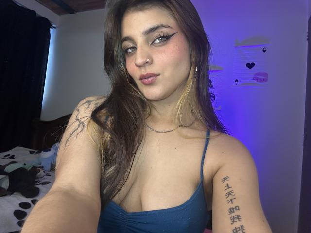 SarahhTanyas with brown hair, blue eyes, and pink lipstick, wearing a blue spaghetti strap top, showing her tits and tattoo on her right arm, in a dimly lit room.