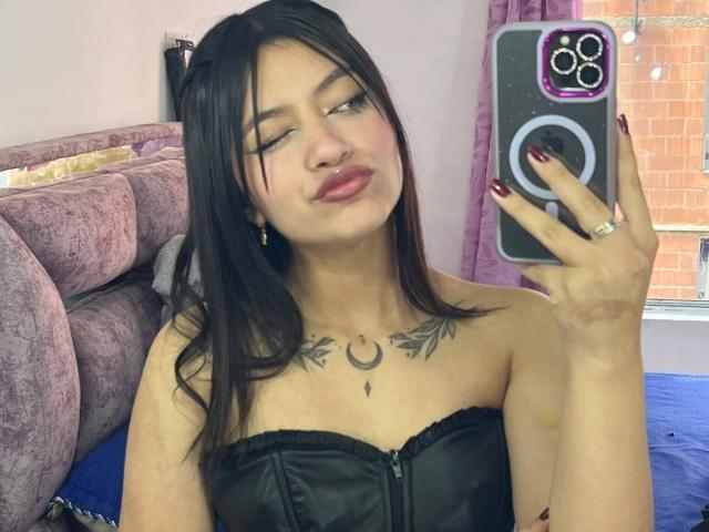ScarlettPreatty with black hair, winking, holding phone, showing off black strapless top, chest tattoo, and ring, with red nails, in a mirror selfie. Boobs visible, looking cute.
