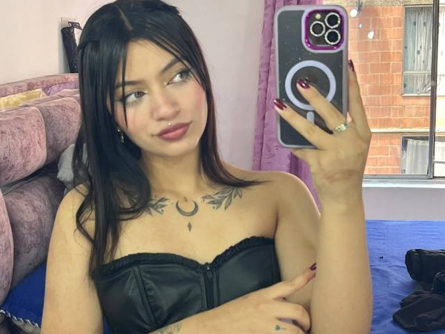 Scarlett, con i capelli neri, il rossetto rosso e una top in pelle nera senza spalline, che mostra il seno e un tatuaggio a forma di mezzaluna, sta scattando un selfie con un anello al dito..