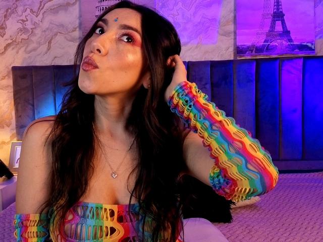 MiaReed with colorful, wavy-patterned arm sleeves and dress, pouting with her hand in her dark hair, showing her tits and a small makeup dot on her forehead.