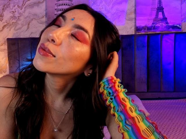 MiaReed with closed eyes, dark hair, and colorful rainbow wristband, has pink eyeshadow and a small blue dot on her forehead. She's touching her head with a relaxed expression.
