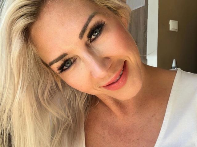 Cindy-Stone1-hot ha i capelli biondi, gli occhi azzurri e un sorriso carino con labbra rosa. Indossa una maglietta bianca che lascia intravedere un po' il seno. Sta guardando la fotocamera..