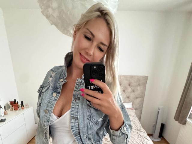 Cindy-Stone1-atractiva, con cabello rubio, vistiendo una chaqueta vaquera y una camiseta blanca, mostrando sus pechos en un selfie frente al espejo, uñas rosadas, sonriendo, sosteniendo un teléfono negro..