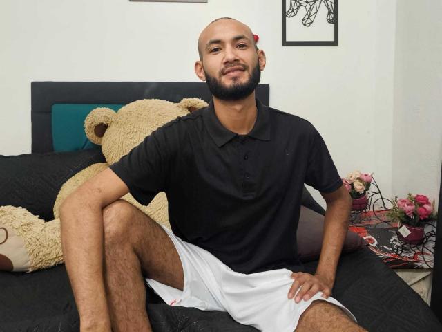 krillynnt with a beard, bald, wearing a black polo and white shorts, sitting on a bed with a giant teddy bear, smiling at the camera.