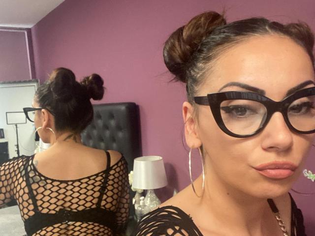 AlexisGray con i capelli castani raccolti in chignon, indossa una maglia nera a rete, ha un sedere grande e un seno prosperoso, porta occhiali neri e orecchini a cerchio, riflessa nello specchio. Sullo sfondo, una parete viola..