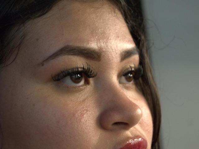 O rosto de Kelly Shibary, com olhos castanhos e longos cílios, lábios rosados levemente entreabertos e um tom de pele claro com sardas sutis. Ela tem cabelo castanho escuro no lado esquerdo..