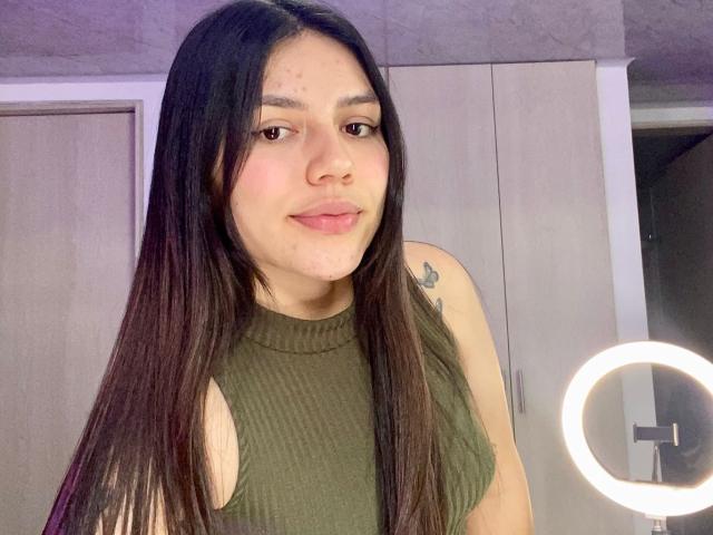 SophiaMarx has long black hair, a cute nose, and a small tattoo on her right shoulder. She's wearing a green ribbed sleeveless top and smiling slightly.
