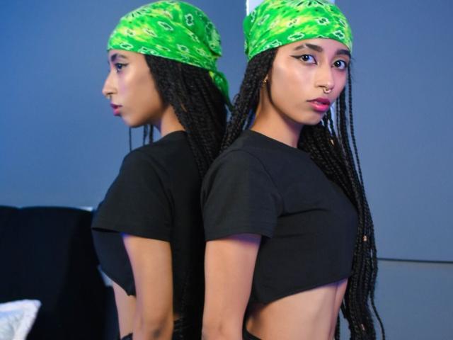 AngelinaStar69, con una bandana verde, un top corto nero e lunghe trecce, è in piedi di fronte a uno specchio. Il riflesso mostra la stessa angolazione, mettendo in risalto il suo anello al naso e il rossetto rosa..