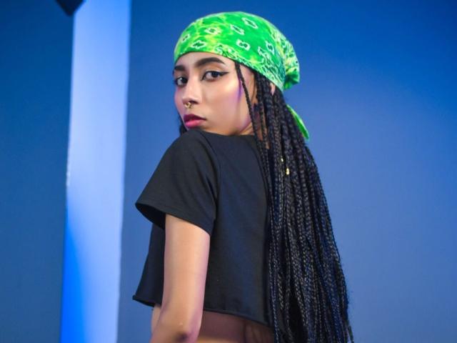 AngelinaStar69 avec de longues tresses noires, un bandana vert, un crop top noir, regardant en arrière, un anneau de nez, fond bleu, expression sérieuse..
