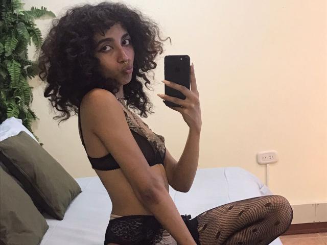 AngelinaStar69 mit lockigem, schwarzem Haar, trägt einen schwarzen Spitzen-BH und Netzstrümpfe, sitzt auf dem Bett und macht ein Selfie mit ihrem Handy, wobei sie ihr Gesäß und ihre kleinen Brüste zeigt..