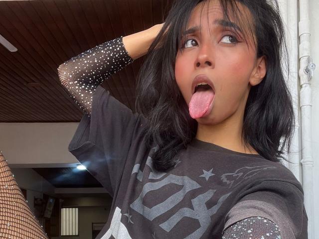 AngelinaStar69, avec ses cheveux noirs, sa peau brune et sa langue tirée, portant une chemise noire à motifs d'étoiles et des manches en résille, se touche la tête. Sa langue est d'un rose bien visible..