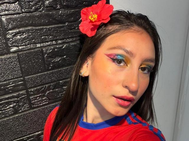 MaddyCarina has long brown hair with a red flower, colorful eyeshadow, and wears a red shirt with blue accents. She has pink lipstick and small earrings.