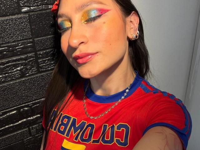 MaddyCarina has colorful, gradient eye makeup with yellow, blue, and orange, wearing a red sports shirt with blue trim, a chain necklace, and a red hair clip.