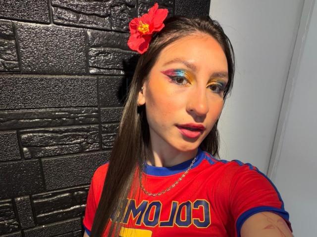MaddyCarina with long brown hair, red flower, yellow and blue eye makeup, red shirt, necklace, standing against black brick wall. Her tits are small, she has a cute face.