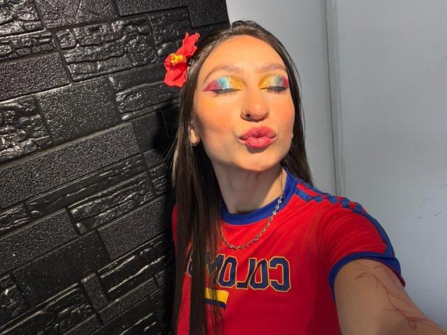 MaddyCarina with colorful eyeshadow, pouty lips, red shirt, and blue stripes, wearing a red flower in her brown hair, leaning against a black textured wall.