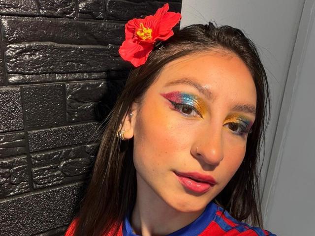 MaddyCarina has bright, colorful eyeshadow with yellow, orange, and blue hues, a red flower in her brown hair, and a small nose ring. She's wearing a red and blue shirt.