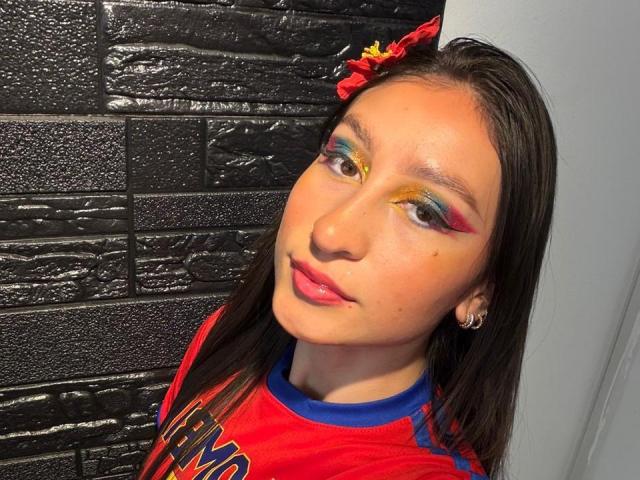 MaddyCarina with colorful eyeshadow, red lips, and hoop earrings, wearing a red sports shirt with blue trim, black textured wall in background. She's looking slightly to the right.