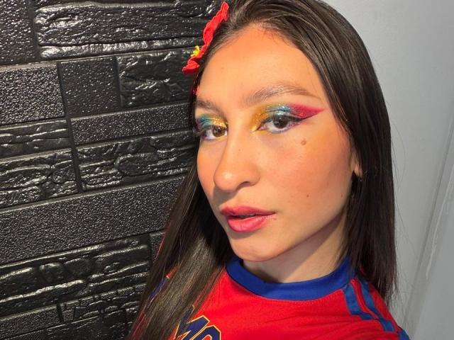 MaddyCarina has long brown hair, yellow-green-blue-orange eye makeup, red lips, and a small mole on her right cheek. She's wearing a red sports shirt with blue trim.