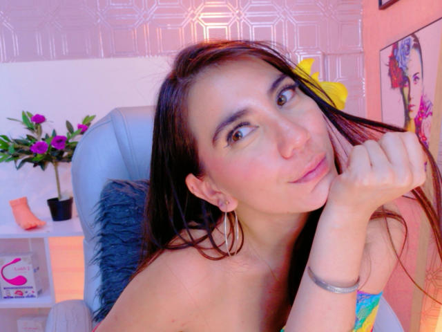 FreyaraVenwo con lunghi capelli castani, sorridente, che si tiene i capelli, pelle rosea, occhi marroni, seno piccolo, senza vestiti, fiore giallo, sedia blu, cuscino grigio, vaso rosa, mensole bianche..