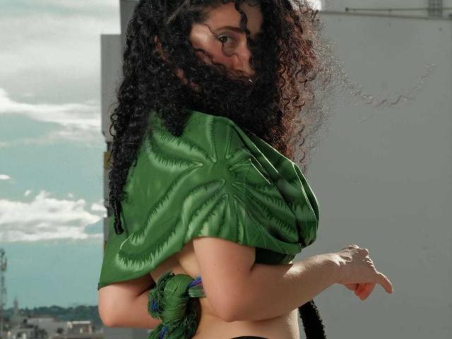 NoaxxMalak with curly black hair, green puffed-sleeve top, exposed tan skin, and green fabric tied around her waist, looking over her shoulder. Boobs partially visible.