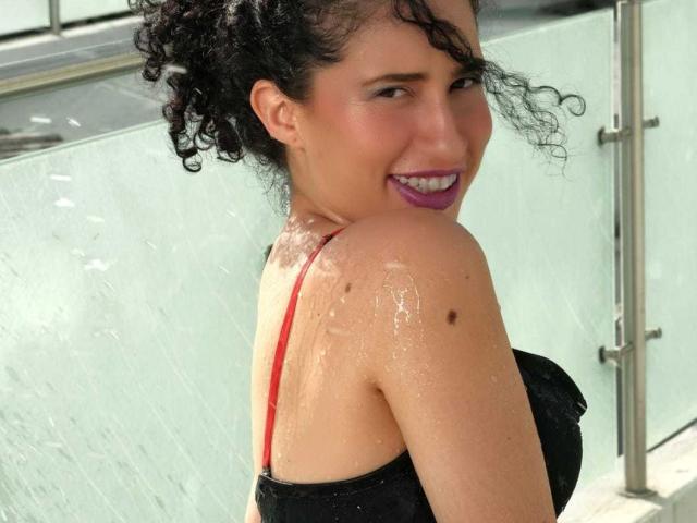 NoaxxMalak, con el pelo negro rizado y mojado, sonriendo con lápiz labial morado, vestida con un top negro y una correa roja, mostrando su espalda y un hombro mojado con una pequeña peca..