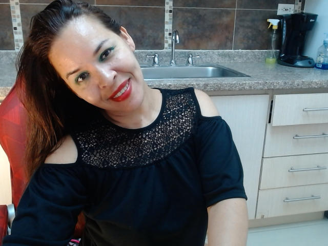 LizethBells, con un lápiz labial rojo brillante, una blusa de encaje negro y hombros descubiertos, sonriendo a la cámara, sentada en una cocina con una encimera de granito y un fregadero de acero inoxidable..