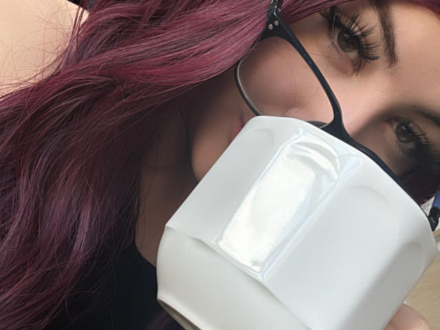 IsabellWinter with purple hair, wearing glasses, and a white face mask, holding the mask to her nose and mouth, showing her dark eyebrows and long eyelashes.