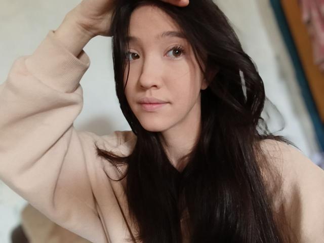 AshleyNaone, com cabelos longos e escuros, olhos castanhos claros e um sorriso discreto, vestindo um suéter bege, levantando a mão em direção à cabeça. Suas nádegas não estão visíveis..