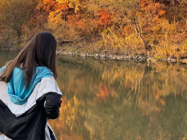 AshleyNaone with long brown hair, wearing a blue and black jacket, stands by a reflective lake, facing autumn trees with orange and yellow leaves.