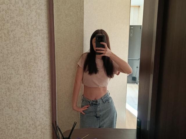 AshleyNaone, con cabello castaño largo, un top rosa corto y jeans azules de cintura alta, está de pie frente a un espejo, con una mano en la cadera y un teléfono frente a ella, cubriendo su rostro..