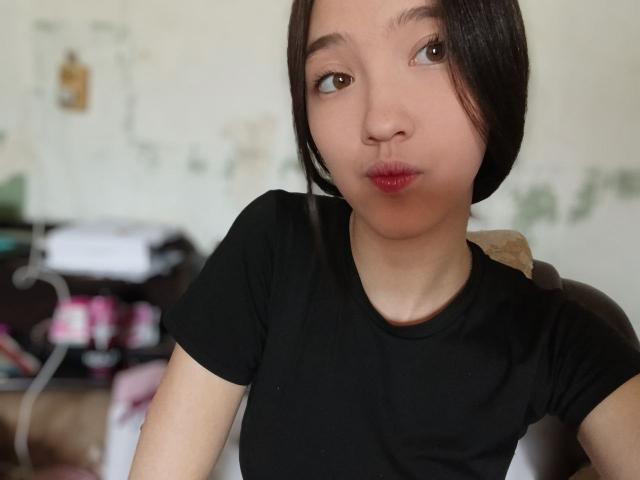 AshleyNaone, avec ses cheveux noirs courts, ses lèvres rouges et pulpeuses et sa peau claire, porte un t-shirt noir et est assise sur une chaise. Ses petites seins sont partiellement visibles..