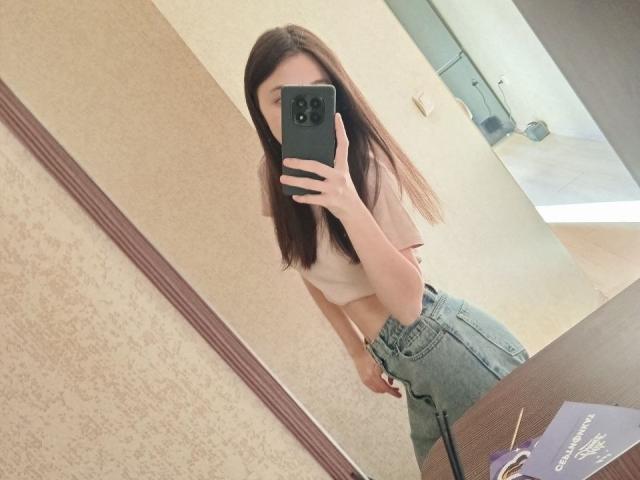 AshleyNaone maakt een foto van zichzelf in de spiegel, met een roze topje, waar haar kleine borsten zichtbaar zijn, en hoge blauwe jeans. Ze houdt een zwarte telefoon vast en heeft lang bruin haar..