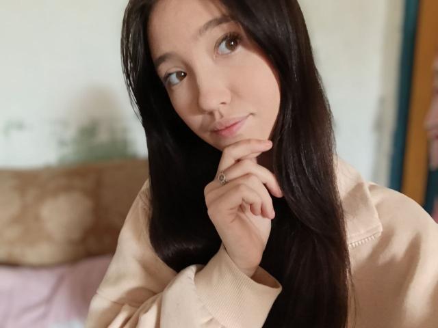 AshleyNaone with long black hair, brown eyes, and a small smile, wearing a beige jacket, touching her chin with her hand, showing a ring. Soft light, relaxed look.