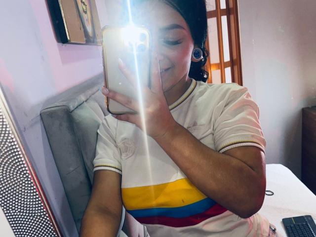 LonxEva with dark hair, medium skin, wearing a white shirt with yellow, red, and blue stripes, taking a mirror selfie, smiling, flash from phone, small earrings, in a room.