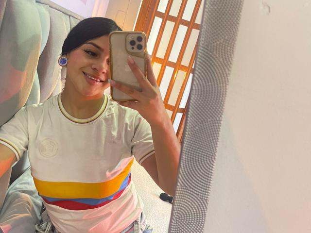 LonxEva with tan skin, black headscarf, white shirt with yellow, red, and blue stripes, holding phone, smiling, earbuds, mirror selfie, carpeted floor, wooden door in background.