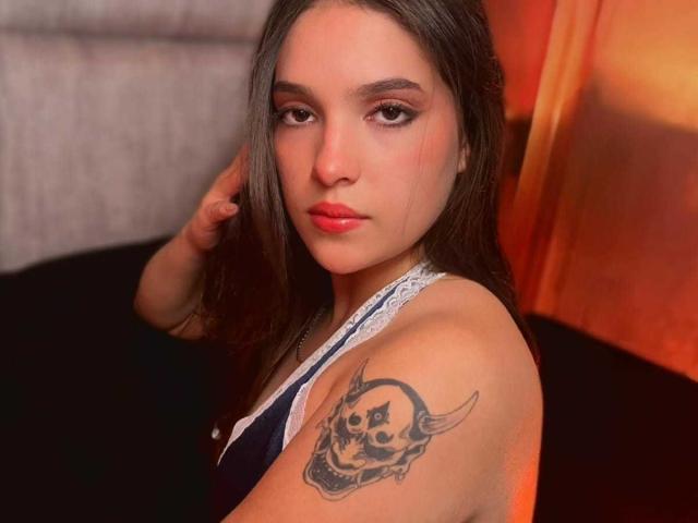 Katarina Novak mit langen, braunen Haaren, rotem Lippenstift und einem Totenkopf-Tattoo auf ihrer rechten Schulter, die in die Kamera lächelt, trägt eine blaue und weiße Spitzenbluse..