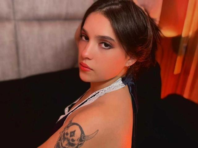 Katarina Novak mit dunkelbraunen Haaren in einem lockeren Dutt, die über ihre Schulter schaut, trägt ein weißes Spitzen-Haltertop und zeigt ein Totenkopf-Tattoo an ihrer rechten Schulter..