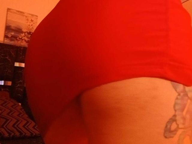 ZarayFeet's butt in tight red shorts, showing a tattoo on the right thigh. Warm, orange lighting. The tattoo is a small, gray, line-drawing style design.