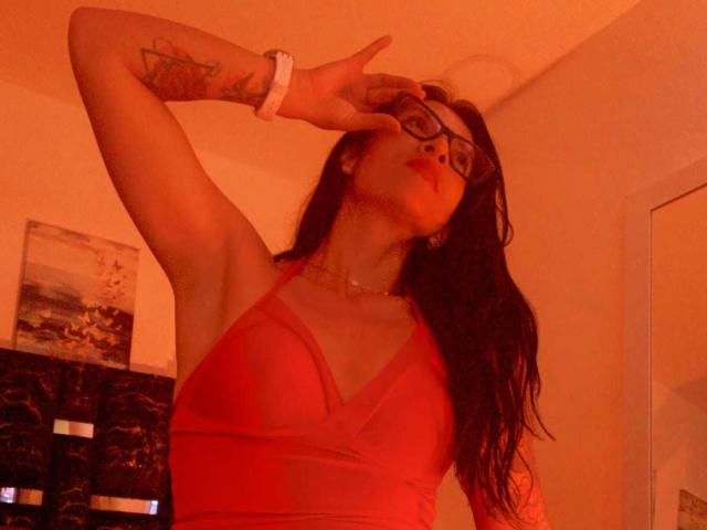 ZarayFeet, con cabello largo y oscuro, vestido rojo, gafas y un tatuaje en el brazo, haciendo el gesto de la paz, con luz roja y una pulsera blanca, de pie en una pose segura y sensual..