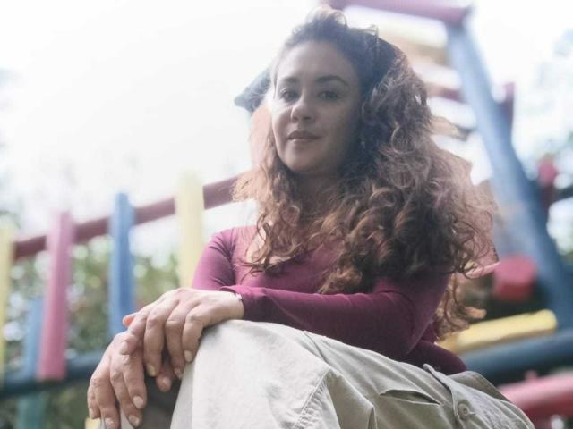 TheresaLarson with curly red hair, wearing a red long-sleeve shirt and green pants, sitting and smiling slightly. Her hands rest on her knee. Soft, natural light.