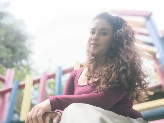 Theresa Larson, com cabelos castanhos cacheados, vestindo uma blusa marrom-avermelhada e uma saia branca, sentada em equipamentos de playground coloridos, sorrindo levemente. Suas mãos estão apoiadas nos joelhos..