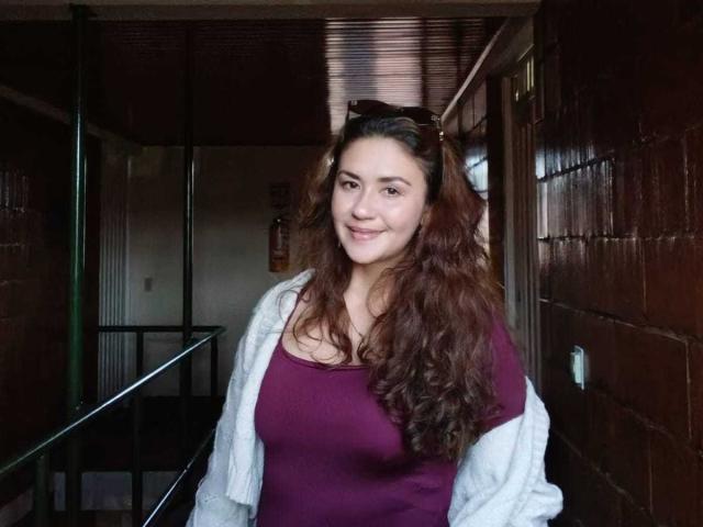 Theresa Larson tem cabelos castanhos longos e ondulados, uma blusa roxa e um cardigã branco, e está em um corredor pouco iluminado, com um sorriso discreto. Seus seios são pequenos..
