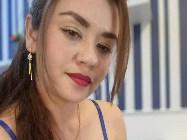 TheresaLarson has light skin, brown hair, and red lipstick, wearing blue straps, gold earrings, and black eyeliner, with a subtle smile, looking down.