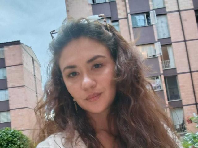 Theresa Larson, con cabello castaño ondulado, piel clara y ojos marrones, vestida con una camisa blanca, está de pie frente a un edificio de apartamentos rosa y marrón, mirando ligeramente hacia un lado..