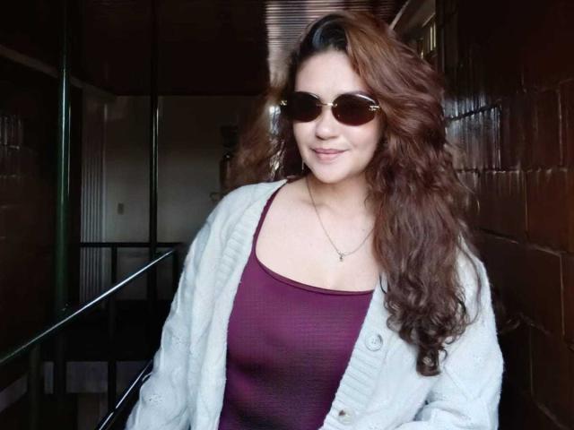 Theresa Larson, con cabello castaño largo y ondulado, usando gafas de sol, un cárdigan blanco y una camiseta morada, que deja ver ligeramente sus pechos. Sonriendo, de pie en el interior..