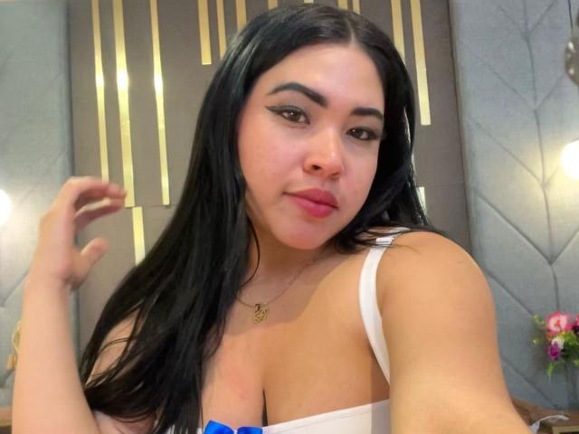 OliviGrace with long black hair, wearing a white top showing her big boobs, has pink lips and a gold necklace, looking at the camera with a slight smile.