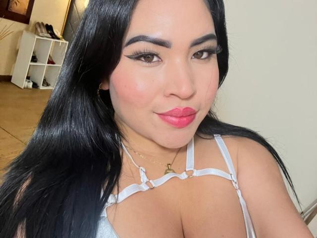 OliviGrace mit langen, schwarzen Haaren, die ein weißes Top mit dünnen Trägern trägt, wobei ihre großen Brüste sichtbar sind, mit rosa Lippenstift und einem leichten Lächeln, steht in einem gut beleuchteten Raum..