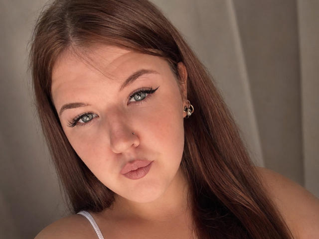 TinaAdie heeft lichtgroene ogen, lang bruin haar, een neusring en draagt een kleine gouden oorbel met een schattig gezicht en lichtjes opgetrokken lippen. Ze kijkt naar de camera..