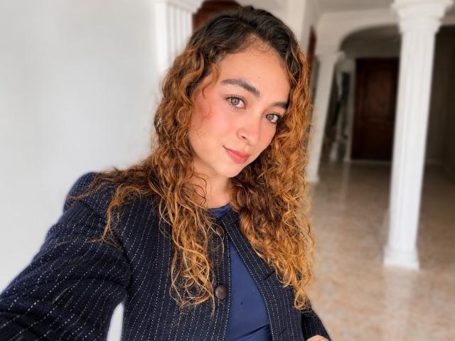 Arianna Blake, con i capelli castano ricci, indossa un cardigan scuro a righe sottili sopra una camicia blu, ha un viso carino e un naso piccolo, ed è in piedi in un corridoio bianco..