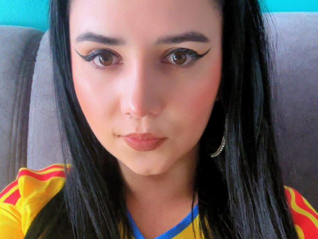 Kkatalina hat schwarze Eyeliner-Akzente, braune Augen und trägt ein gelb-rotes Hemd mit einem blauen Ausschnitt. Sie hat Ohrringe im Hula-Hoop-Stil und schaut in die Kamera..
